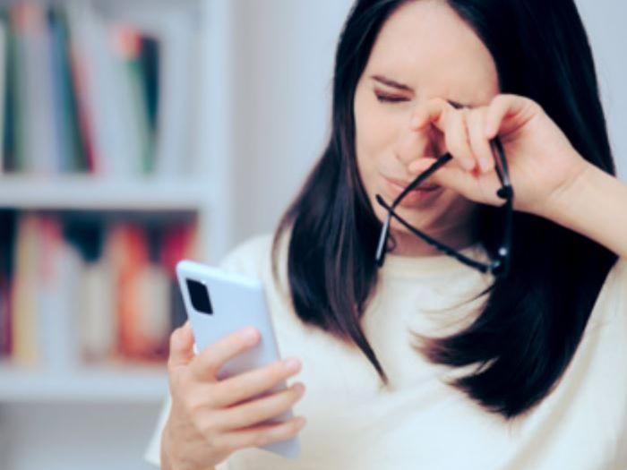 Zdjęcie przedstawiające kobietę w wieku powyżej 30 lat, przecierającą oczy podczas spoglądania na ekran swojego telefonu.