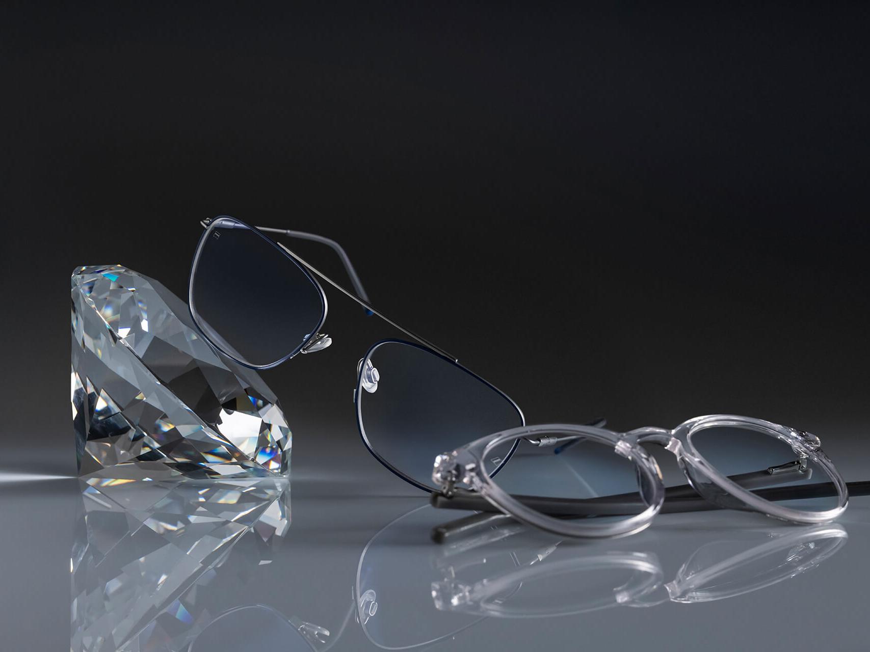 Dwie pary okularów z soczewkami ZEISS, które posiadają najtwardsze dostępne powłoki ZEISS – DuraVision® Platinum. Jedna para okularów jest oparta o kryształ, druga leży płasko na ziemi. Obie posiadają przejrzyste soczewki bez niebieskich refleksów.