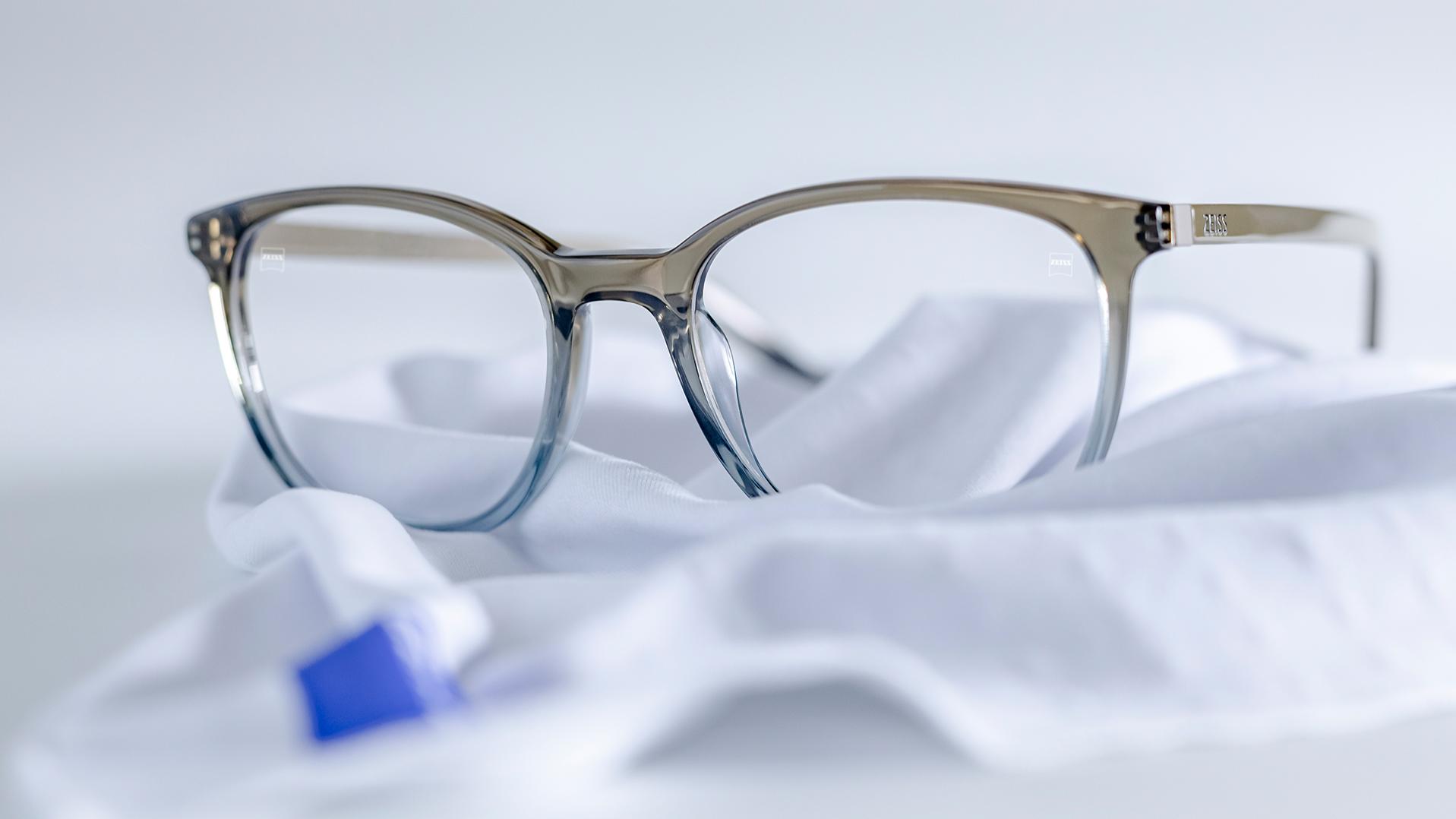 Para okularów z szaro-niebieskimi oprawkami i soczewkami ZEISS z powłoką DuraVision® leży na białej ściereczce z mikrofibry.
