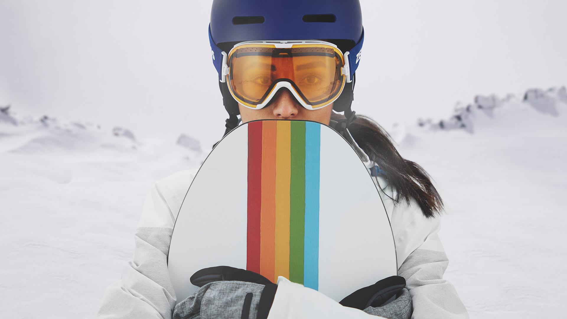 Młoda kobieta nosząca gogle narciarskie z technologią ZEISS Interchangeable trzyma deskę snowboardową.