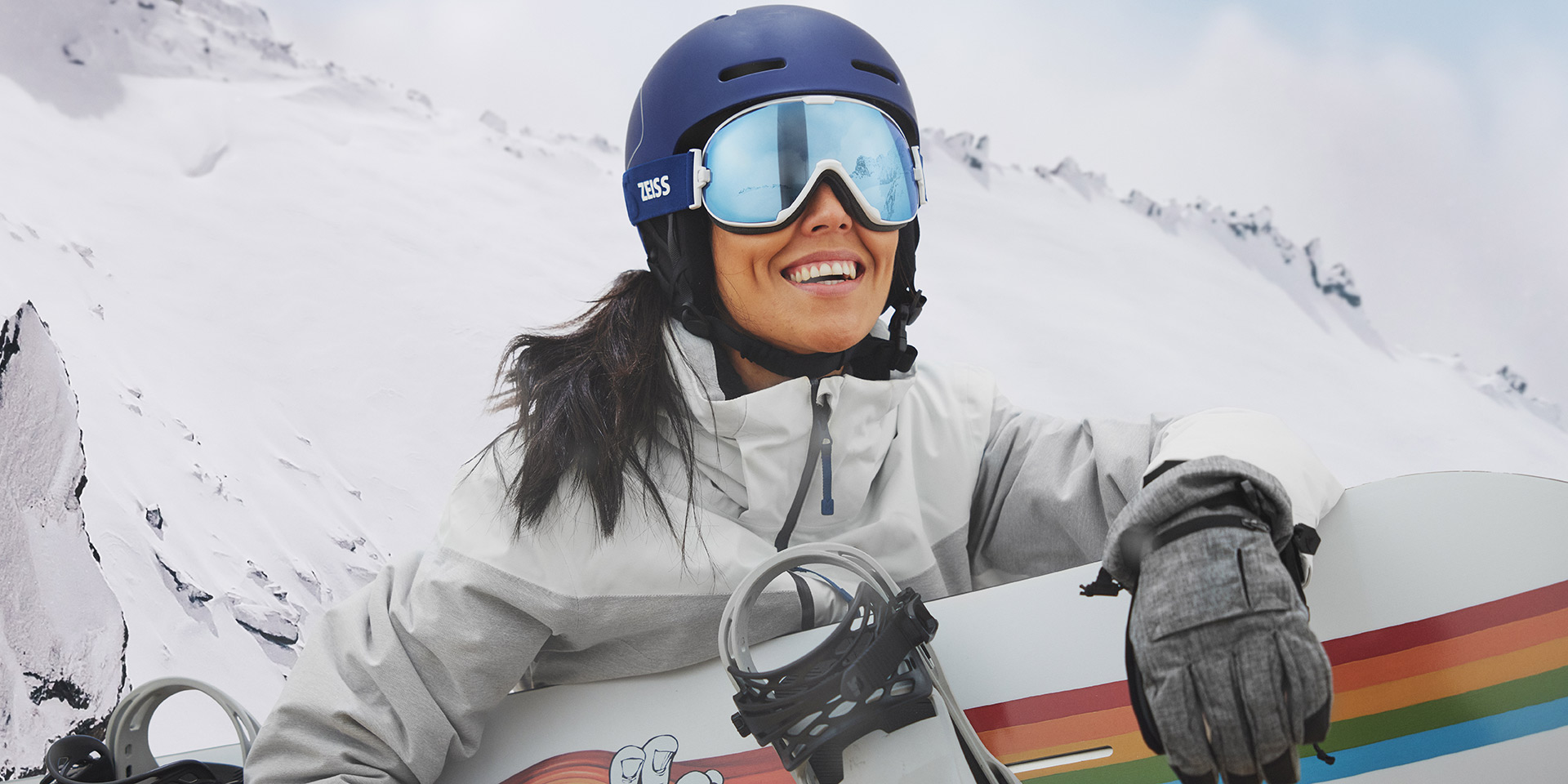Młoda kobieta nosząca gogle narciarskie z technologią ZEISS Interchangeable trzyma deskę snowboardową.