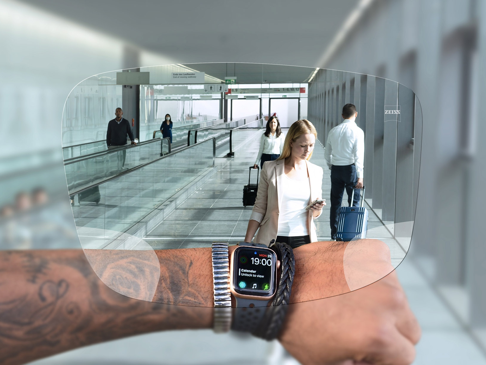 Osoba spogląda na zegarek przez okulary z soczewkami ZEISS SmartLife Digital. W tle widać typową sytuację na lotnisku z ludźmi pchającymi walizki, przemieszczającymi się na ruchomych schodach. 